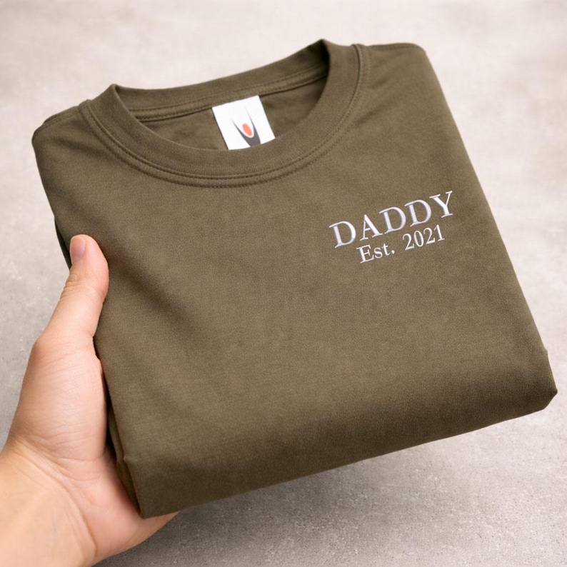 May include: Olive green crew neck t-shirt with the white text "DADDY Est. 2021" printed on the front. The shirt is folded neatly, showcasing the design. The shirt is made of a soft material.