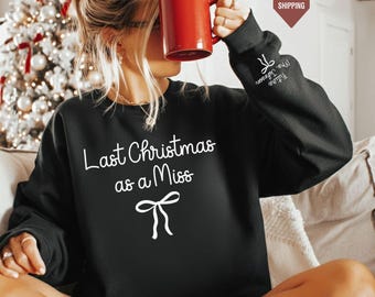 Könnte beinhalten: Ein schwarzes Langarm-Sweatshirt mit dem weißen Schriftzug "Last Christmas as a Miss" über einer weißen Schleifengrafik. Der linke Ärmel zeigt den weißen Text "Future Mrs. Johnson" mit einer kleinen Schleife. Eine Person hält einen roten Becher.