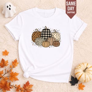 May include: A white short-sleeved t-shirt featuring a central graphic of a cluster of pumpkins with various patterns including leopard print, black and white buffalo plaid, polka dots, and solid brown, accented with small stars and swirls.