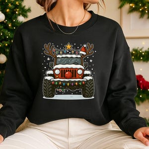 Puede incluir: Sudadera negra con una ilustración de dibujos animados de un Jeep rojo decorado para Navidad. El Jeep tiene cuernos de reno, un gorro de Papá Noel y una nariz roja. También está decorado con luces navideñas y nieve.