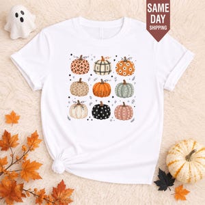 May include: A white short-sleeve t-shirt featuring a graphic print of nine pumpkins in various patterns and colours, including leopard print, plaid, floral, and polka dot designs, arranged in a three-by-three grid with small star and leaf accents.