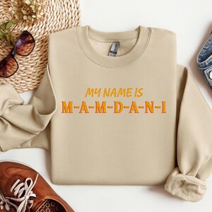 May include: A beige sweatshirt with the text "MY NAME IS M-A-M-D-A-N-I" in orange and yellow lettering. The sweatshirt is displayed with brown shoes, sunglasses, and denim jeans.
