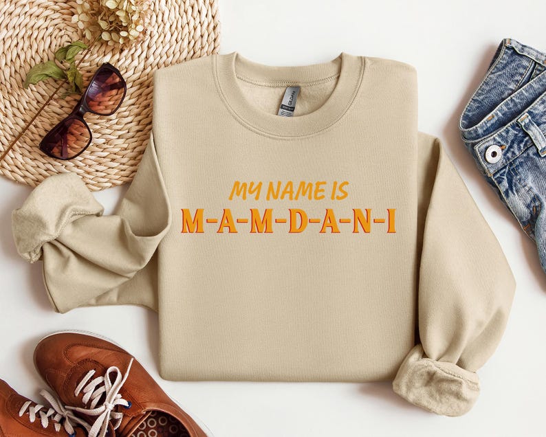 May include: A beige sweatshirt with the text "MY NAME IS M-A-M-D-A-N-I" in orange and yellow lettering. The sweatshirt is displayed with brown shoes, sunglasses, and denim jeans.