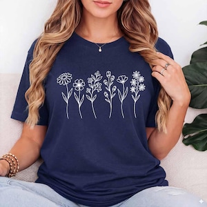 May include: A navy blue short-sleeved t-shirt featuring a white line art graphic of six distinct wildflowers arranged in a horizontal row across the chest. The design includes various stems, leaves, and blossoms, creating a botanical pattern.