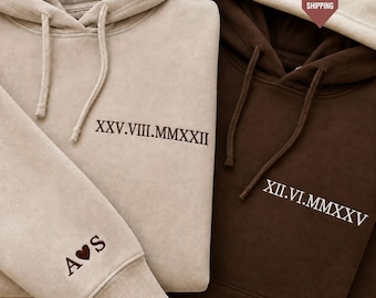 Könnte beinhalten: Ein beigefarbener Kapuzenpullover mit dunkelbrauner Stickerei XXV.VIII.MMXXII und einem A Herz S Detail am Ärmel, neben einem dunkelbraunen Kapuzenpullover mit weißer Stickerei XII.VI.MMXXV. Beide Pullover haben passende Kordelzüge und Kapuzen.