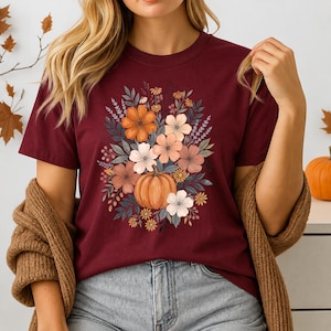 以下が含まれることがあります： ボルドー色の半袖Tシャツ。中央にはカボチャと、オレンジ、ピンク、白、くすんだ緑色の花や葉が描かれたグラフィックプリントがあり、茶色のニットカーディガンを重ね着しています。