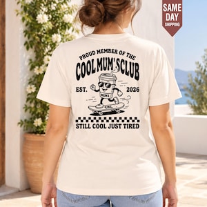 Peut inclure: Un t-shirt couleur crème avec un motif noir au dos. Le design représente une tasse de café sur un skateboard avec le texte PROUD MEMBER OF THE COOL MUM'S CLUB EST. 2026 et STILL COOL JUST TIRED, accompagné d'une bordure à damier.