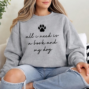 May include: A gray sweatshirt with the text "all I need is a book and my dog" and a paw print graphic. The sweatshirt is paired with blue jeans. The overall theme is casual wear.