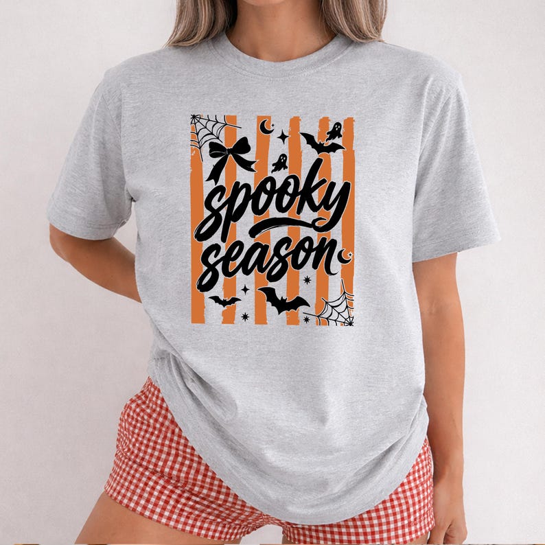 May include: A light grey short-sleeve t-shirt featuring a central graphic with vertical orange stripes and black text that reads spooky season. The design includes black illustrations of spiderwebs, a bow, bats, ghosts, and stars.