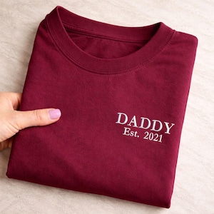 May include: A folded burgundy t-shirt with the word "DADDY" and "Est. 2021" embroidered in white. The shirt is made of a soft material and has a crew neck.