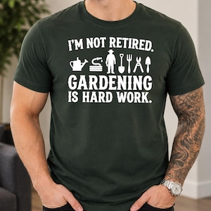 May include: A dark green short-sleeved t-shirt featuring white text that reads I'M NOT RETIRED. GARDENING IS HARD WORK. The design includes white graphic icons of a watering can, a coiled hose, a person, a shovel, a pitchfork, shears, and a trowel.
