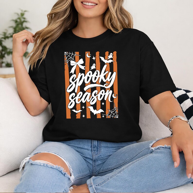 May include: A black t-shirt featuring a graphic print with vertical orange and black stripes. The design includes the white text "spooky season" in a script font, surrounded by white illustrations of a bow, spiderwebs, bats, and stars.