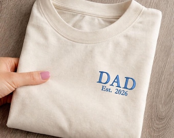 Könnte beinhalten: Cremefarbenes T-Shirt mit dem Wort "DAD" in Blau gestickt, zusammen mit "Est. 2026". Das Shirt ist ordentlich gefaltet und zeigt das Design. Das T-Shirt besteht aus einem weichen Material.