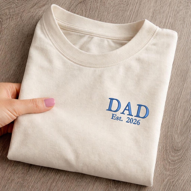 May include: Cream-colored t-shirt with the word "DAD" embroidered in blue, along with "Est. 2026". The shirt is folded neatly, showcasing the design. The t-shirt is made of a soft material.
