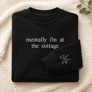 May include: Black sweatshirt with white embroidered text reading "mentally i'm at the cottage". The sleeve displays a golf-themed emblem with the numbers "24" and "81" in red and blue.