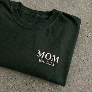 May include: Dark green t-shirt with the word "MOM" embroidered in white, along with "Est. 2023". The shirt is folded, showcasing the text on the front. The fabric appears soft and comfortable.