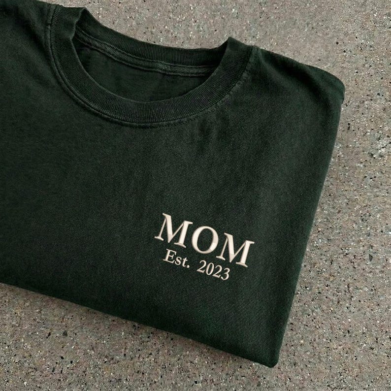 May include: Dark green t-shirt with the word "MOM" embroidered in white, along with "Est. 2023". The shirt is folded, showcasing the text on the front. The fabric appears soft and comfortable.