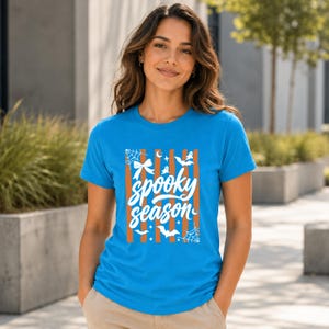 May include: A bright blue short-sleeved t-shirt featuring a central graphic with vertical orange stripes and white text that reads "spooky season." The design includes white illustrations of spider webs, bows, bats, stars, and crescent moons.