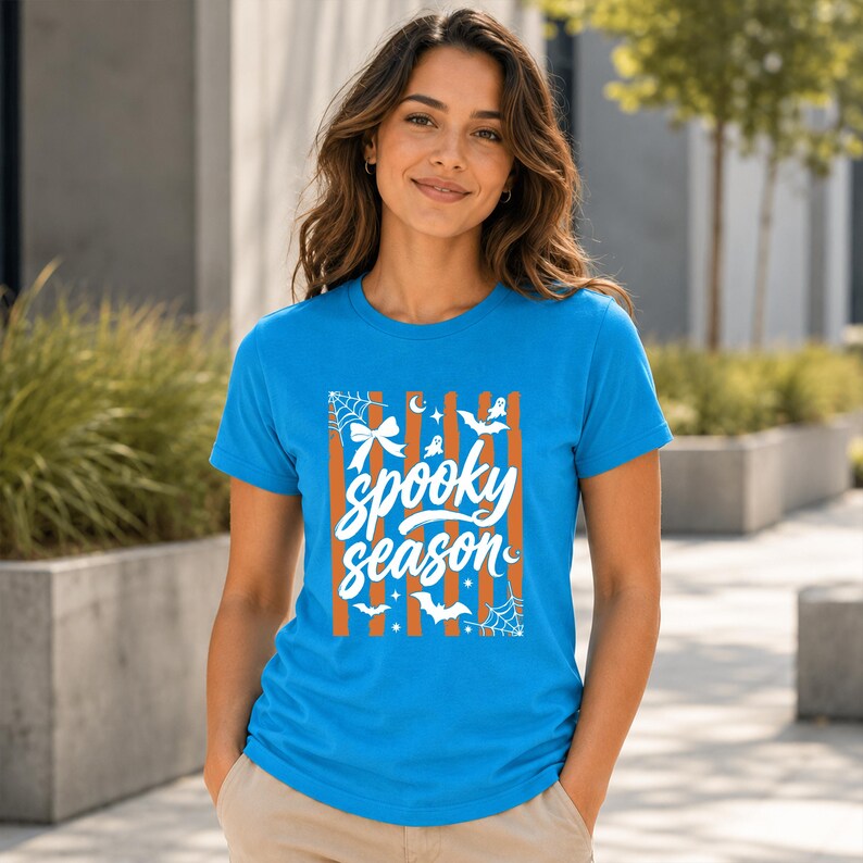 May include: A bright blue short-sleeved t-shirt featuring a central graphic with vertical orange stripes and white text that reads "spooky season." The design includes white illustrations of spider webs, bows, bats, stars, and crescent moons.