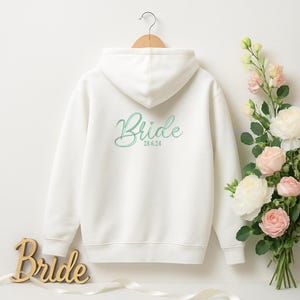 May include: A white hoodie with the word "Bride" embroidered in a cursive font on the back, and the date "28.6.24". The sweatshirt is hanging on a wooden hanger. A wooden sign that says "Bride" is in the foreground.