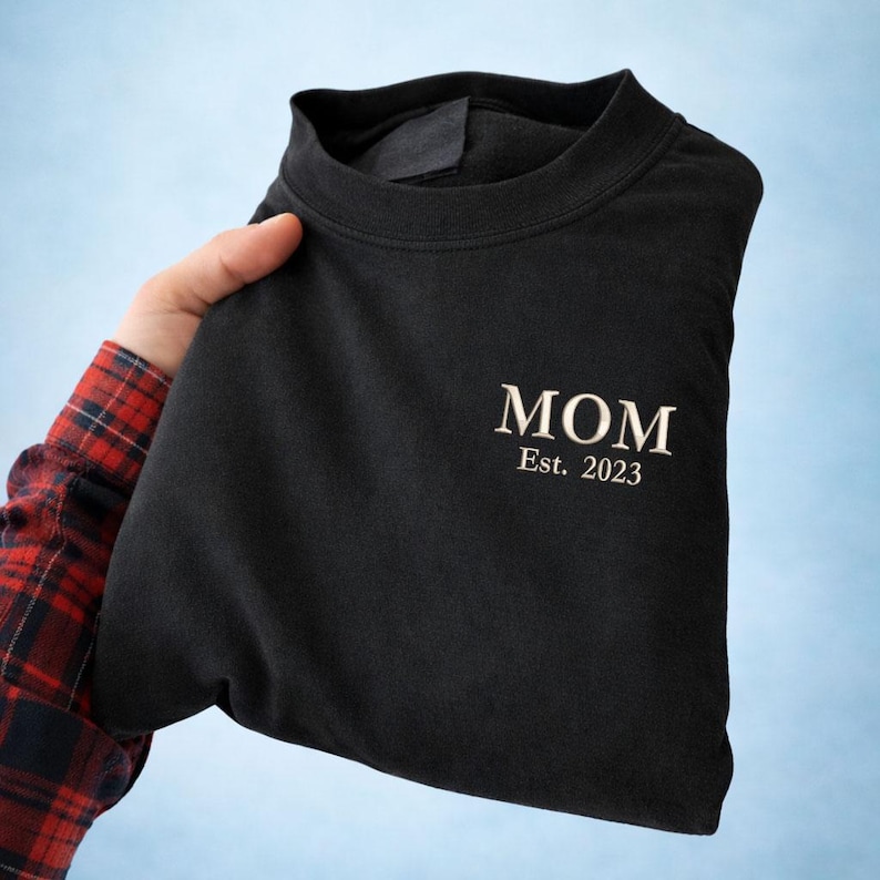 May include: Black crew neck t-shirt with the word "MOM" and "Est. 2023" embroidered in a light color. The shirt is folded and held by a person wearing a red and black plaid shirt.