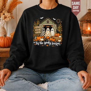 May include: A black crewneck sweatshirt featuring a graphic print of three ghosts standing in front of a barn with pumpkins, corn stalks, and a crow. The text reads 'Tis the Spooky Season' with small orange hearts and white bats in the background.
