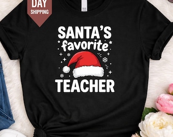 Könnte beinhalten: Ein schwarzes Kurzarm-T-Shirt mit dem weißen Text SANTA'S favorite TEACHER und einer Grafik einer rot-weißen Weihnachtsmütze in der Mitte, umgeben von kleinen weißen Stern- und Schneeflocken-Akzenten, die auf die Vorderseite gedruckt sind.