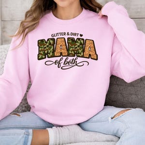 May include: A pale pink sweatshirt featuring the text "Glitter & Dirt Mama of both". The word "Mama" is designed with camouflage and leopard print. The sweatshirt is a long-sleeved crew neck.