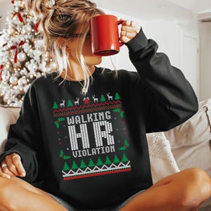 Peut inclure: Sweat-shirt noir avec un motif festif. Le texte "WALKING HR VIOLATION" est affiché en lettres blanches, entouré d'arbres de Noël, de rennes et de houx. Une tasse rouge est tenue devant le visage.