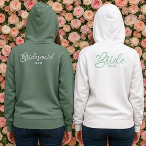 May include: Two hooded sweatshirts, one sage green and one white, with the backs facing the viewer. The green sweatshirt says "Bridesmaid" in cursive with the date "28.6.24" below. The white sweatshirt says "Bride" in cursive with the date "28.6.24" below.