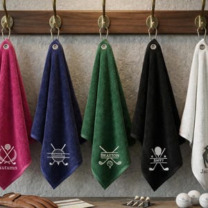May include: Five golf towels in different colours, suspended from a wooden rack. Each towel is personalised with a name and golf-themed graphic. Colours include pink, navy, green, black, and white. Golf balls and a glove are visible below.