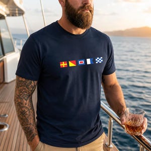 Puede incluir: Una camiseta azul marino de manga corta con una fila horizontal de cinco banderas náuticas de señales impresas en el pecho. Las banderas incluyen una cruz amarilla sobre rojo, un triángulo amarillo y rojo, un cuadrado rojo sobre azul, una bandera blanca y azul, y una bandera a cuadros azul y blanca.