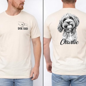 May include: A cream-coloured short-sleeve t-shirt featuring a black graphic on the front chest of a human fist bumping a dog paw with the text DOG DAD. The back displays a detailed black and white sketch of a dog with the name Charlie below it.