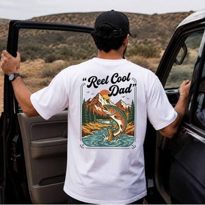 Puede incluir: Una camiseta blanca de manga corta con un gráfico en la espalda que incluye el texto "Reel Cool Dad" sobre la ilustración de un pez saltando en un río, rodeado de pinos, montañas y un sol, enmarcado por dos anzuelos de pesca.