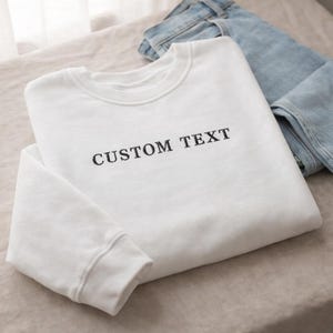 May include: A white sweatshirt with the text "CUSTOM TEXT" in black lettering is folded on a neutral surface. Light blue jeans are partially visible in the background, suggesting a casual outfit.