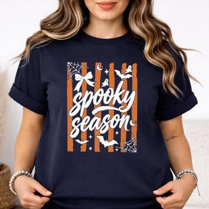 May include: A dark navy blue short-sleeve t-shirt featuring a central graphic with vertical orange and navy stripes. The design includes white text reading "spooky season" surrounded by illustrations of a bow, spiderwebs, bats, and ghosts.