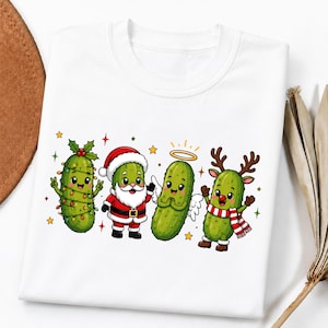 May include: A white t-shirt featuring a graphic print of four cartoon pickles dressed for the holidays. One pickle is wrapped in string lights, one wears a Santa suit, one has an angel halo and wings, and one wears reindeer antlers and a scarf.