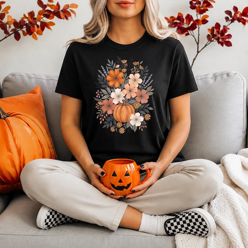 以下が含まれることがあります： 黒の半袖Tシャツ。中央にはオレンジ色のカボチャと、ピーチ、クリーム、落ち着いた緑の色合いの秋らしい花や葉のブーケが描かれています。明るい色のズボンとスニーカーを合わせています。