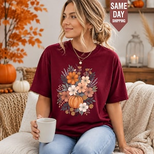 May include: A maroon short-sleeve t-shirt featuring a central graphic print of a pumpkin surrounded by various autumn-colored flowers and foliage in shades of orange, cream, and brown, with a crew neckline and a relaxed fit.