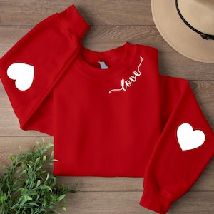 Può includere: Una felpa rossa con cuori bianchi sulle maniche e la parola "love" in corsivo. La felpa è esposta su una superficie di legno, con un cappello e del verde sullo sfondo.