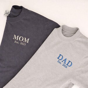 May include: Two crew neck t-shirts. One is dark gray with "MOM Est. 2023" embroidered in white. The other is light gray with "DAD Est. 2026" embroidered in blue. Both shirts are made of a soft material.