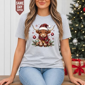 Może przedstawiać: Jasnoszary t-shirt z krótkim rękawem z grafiką przedstawiającą szkocką wyżynną krowę w czerwono-białej czapce Mikołaja i szaliku w kratę, otoczoną bombkami, gałązkami sosny i płatkami śniegu, z czerwonym banerem o treści SAME DAY SHIPPING.