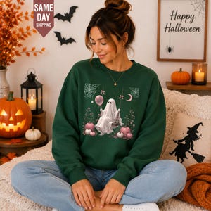 May include: A dark green crewneck sweatshirt featuring a graphic print of a white ghost sitting on a swing, surrounded by pink balloons, spiderwebs, and floral elements. The ghost is draped in a white sheet, and the design includes small bows.