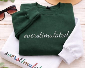 Könnte beinhalten: Zwei Sweatshirts, eines in Dunkelgrün und eines in Weiß, sind gestapelt. Das grüne Sweatshirt hat das Wort "overstimulated" in Weiß gestickt. Das weiße Sweatshirt hat dasselbe Wort in Regenbogenfarben gestickt. Sonnenbrille und Hut im Hintergrund.
