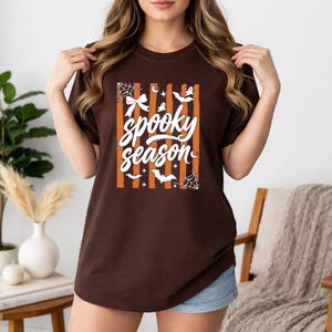 May include: A dark brown short-sleeve t-shirt featuring a central graphic with vertical orange stripes and the white text "spooky season". The design includes white illustrations of a bow, spiderwebs, bats, stars, and small ghosts.