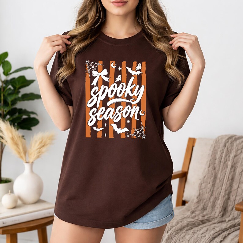 May include: A dark brown short-sleeve t-shirt featuring a central graphic with vertical orange stripes and the white text "spooky season". The design includes white illustrations of a bow, spiderwebs, bats, stars, and small ghosts.