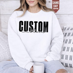 May include: A white long-sleeve crewneck sweatshirt featuring the word CUSTOM in a bold, black, sans-serif font, with the word custom written underneath in a smaller, black, cursive script font. The garment has a ribbed collar and cuffs.