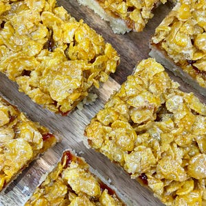 May include: Close-up of several golden-brown cereal bars with a crispy, glazed topping. The bars are cut into rectangular shapes and sit on a wooden surface. The cereal topping is made of corn flakes.