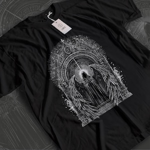Könnte beinhalten: Schwarzes T-Shirt mit einem detaillierten weißen Grafikdesign einer bekleideten Figur mit großen Flügeln, eingebettet in einen Bogenrahmen. Das Design enthält komplizierte Details und einen Halo-Effekt. Ein Etikett zeigt die Größe "LARGE".
