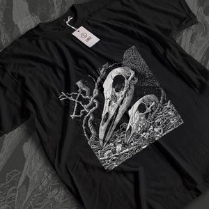 Könnte beinhalten: Schwarzes T-Shirt mit einer detaillierten weißen Grafik von zwei Vogelschädeln und komplizierten, astähnlichen Designs. Das Shirt hat ein Etikett mit der Aufschrift "LARGE". Das Design ist mittig auf der Vorderseite des Shirts platziert.